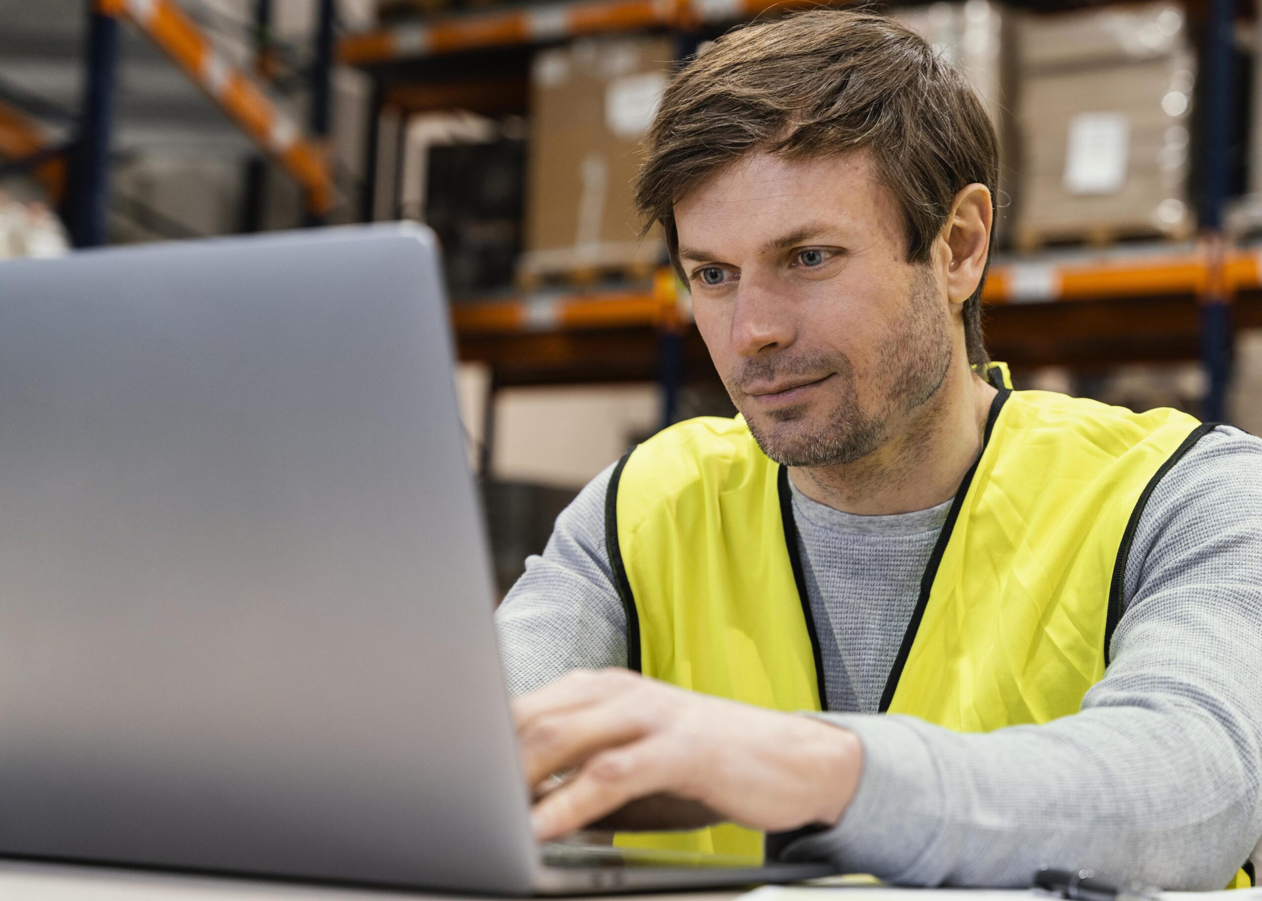 Hombre con chaleco de seguridad en un almacén estudiando con un portátil el máster en logística online